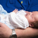 dad blue tshirt, holding baby dressed in white, close up image