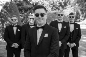 groom, shades, sunglasses, dark specs, black tie, bland white, groomsmen background, smiling, gather