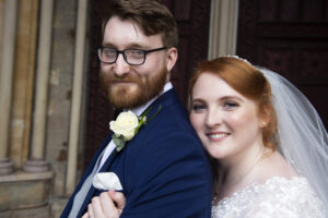 bride red hair, ginger hair, pinned up, hair tied back, white veil, white lace dress, standing behind groom, he smiles, beard, spectacles, glasses, button hold, sideways pose, smiling,