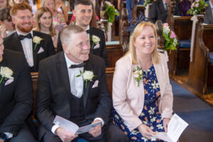 interior church, mother of the bride, pink jacket, blue floral dress, father of the bride, black tie, button hole, smiling guests
