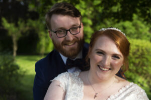 bride foreground, groom background, smiling, close up, eye contact camera
