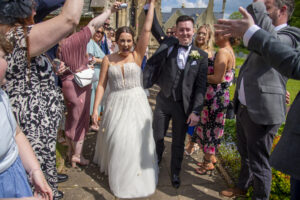 Bride wears white dress, groom wears black tie, guests cheering, people waving, church background