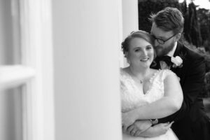 black and white image, groom hugs wife, she is in front, arms around waist, his head nestled in her hair