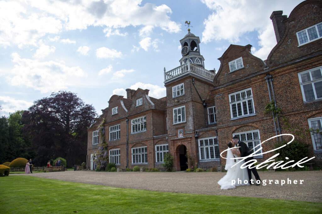 exterior rothamstead manor, blue cloudy sky, gravel drive, green grass, bride and groom right of image, she wears white bridal gown and he wears black tie. kissing