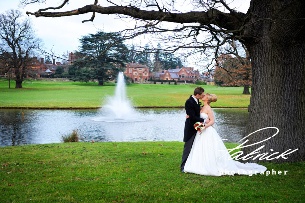 Wedding Hanbury Manor. manor in the background surrounded by trees. foreground the bride and groom kiss. Red and white bouquet. they stand in front of lake and fountain