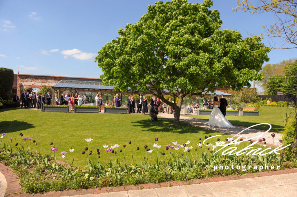 exterior gaynes park, bride and groom walk towards ceremony of guests. trees flowers line the garden