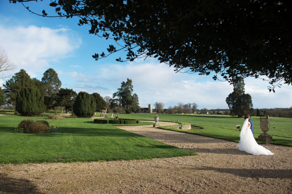 exterior shot of gosfield hall, bride and groom walking up the gravel path, green gardens